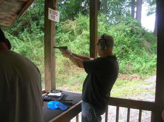 Man in ear protection fires a pistol at an outdoor shooting range, with a "NO RAPID FIRE" sign visible