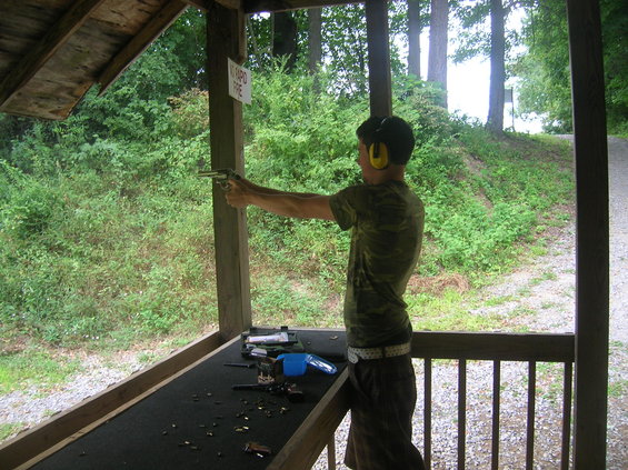 Person wearing ear protection aims a handgun at an outdoor shooting range. Several firearms and shell casings are on the table