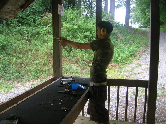 Person in camouflage shirt and shorts at shooting range, aiming a handgun