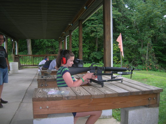Girl with headphones aims rifle at shooting range. Another person stands to the left