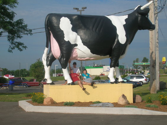 Giant black and white cow statue with three people sitting on its base. Gas station and McDonald's in background