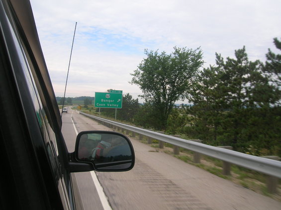 Car interior with side mirror reflecting a person, driving on a highway with a green exit sign for Bangor and Coon Valley