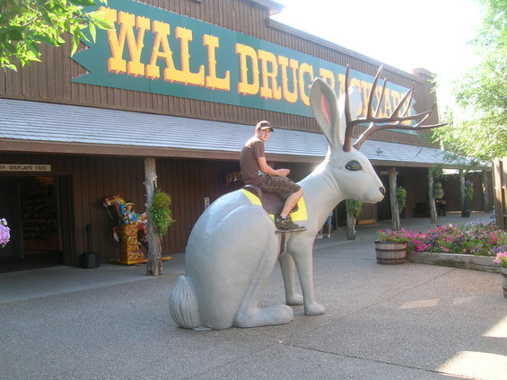 Man sits on a large gray jackalope statue in front of a building with a "Wall Drug" sign
