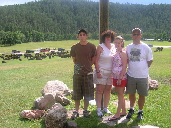 Family of four stands on grass with rocks in foreground; herd of bison graze in background near a road with cars