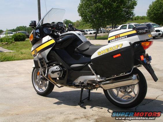 Sheriff's motorcycle with saddlebags and emergency markings parked on pavement, other vehicles in background