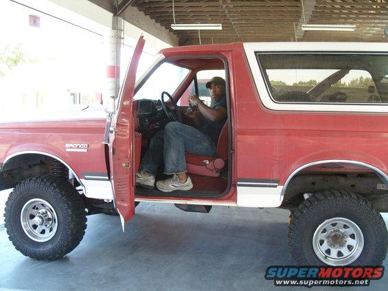Red Ford Bronco with white trim, driver's door open, man in driver's seat. Visible text 'BRONCO' on fender