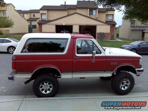 Side view of a red and white 1989 Ford Bronco with a white camper shell and aftermarket wheels