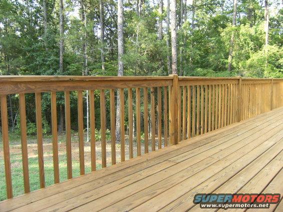 Wooden deck railing and floorboards with trees in the background. Watermark reads SUPERMOTORS.NET