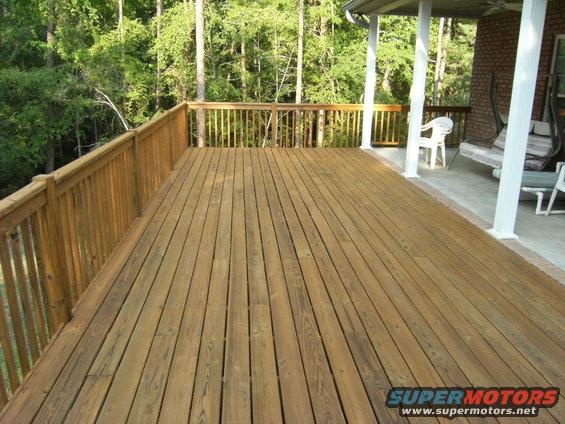 Wooden deck with railing, porch with columns, swing, and chairs. Trees in background