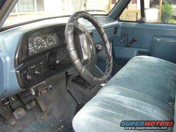 Blue velour bench seat and dashboard of a truck, with a cracked steering wheel cover and floor pedals visible