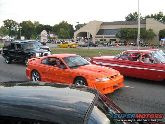 Orange Ford Mustang with flame decals driving in a parade, next to a red classic car