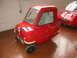 1985 Ford Bronco - A small red microcar is parked indoors next to another vehicle. The license plate is visible.
