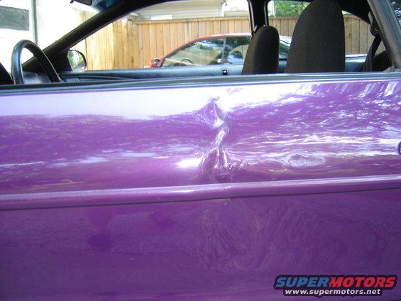 Purple car door with a significant dent near the handle. Another car is visible in the background