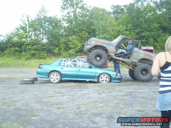 Lifted off-road truck crushing a teal sedan. A person is in the driver's seat of the truck