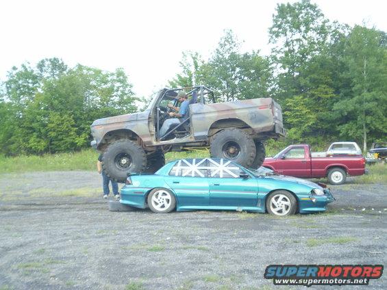 1979 Ford Bronco lifted high on top of a teal sedan, with a red pickup truck behind them