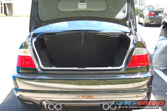 Black BMW sedan trunk open, showing interior. Dual exhaust pipes visible. Another BMW is parked behind