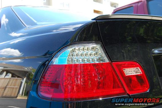 Close-up of a black car's rear LED taillight assembly with a red brake light section and a blue turn signal indicator. Another vehicle is partially visible behind it