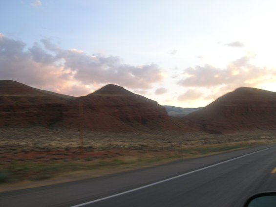 Roadway with white line, passing red-brown hills under a cloudy sky at sunset