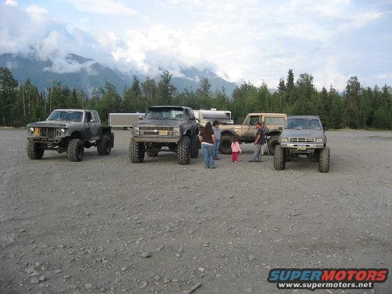 Four lifted trucks and an SUV parked on gravel with mountains in the background