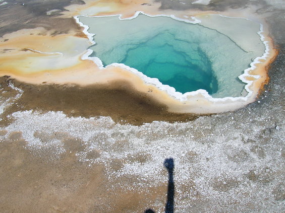 Shadow of a person holding a camera over a hot spring with turquoise water and mineral deposits