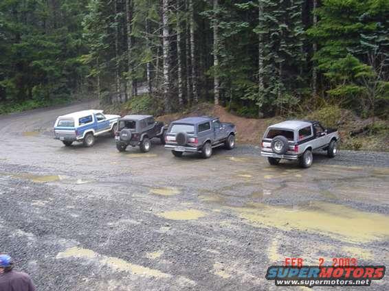 Four SUVs parked on a gravel road in a forest; one is a 1989 Ford Bronco