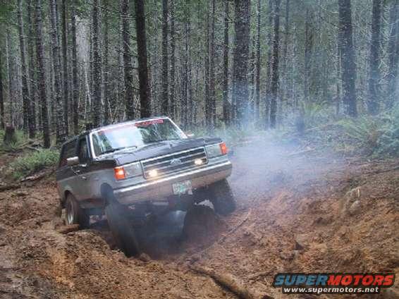 Ford Bronco driving through a muddy forest trail, kicking up smoke