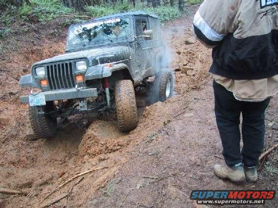 Mud-covered Jeep Wrangler struggling on a muddy trail, front-left tire deep in mud