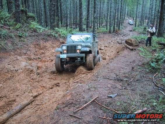 Green Jeep Wrangler navigating a muddy forest trail, with another vehicle in the distance
