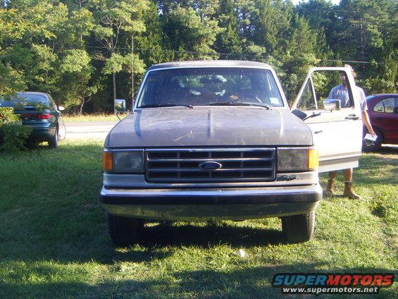 Front view of a gray 1987 Ford F-150 pickup truck with the driver's door open; another car is visible to the left