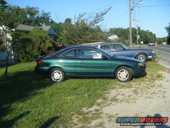Green coupe with a "For Sale" sign in the window, parked next to a blue sedan