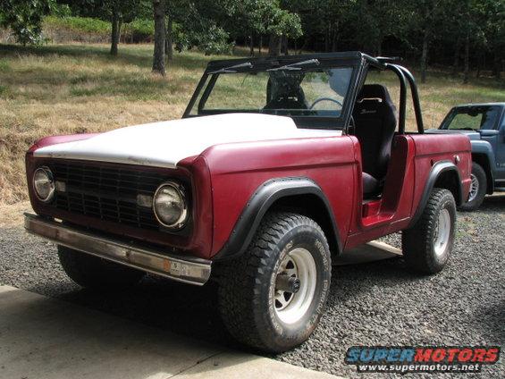 Red 1968 Ford Bronco with a white hood and black roll cage parked on gravel