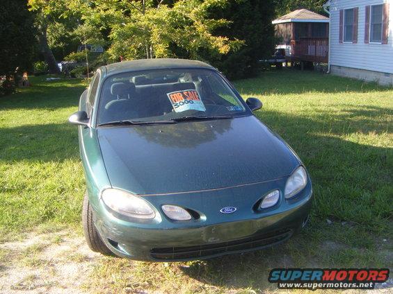 Green Ford Escort with a 'For Sale $3000' sign in the windshield parked in a grassy yard