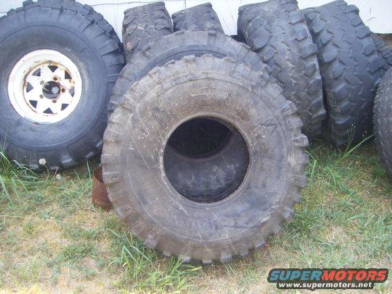 Pile of large, used off-road tires, one with a white rim, on grass next to a building
