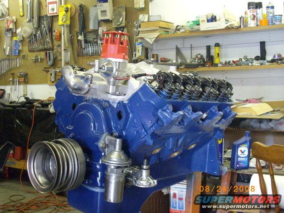 Blue V8 engine block with red distributor cap, fuel pump, and pulley system. Garage background with tools and shelves