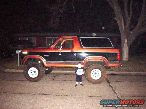 Side view of a lifted 1980s Ford Bronco, orange and black two-tone paint, with a child standing by the rear tire