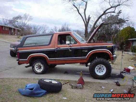 Two-tone orange and black Ford Bronco with hood open, jacked up with a spare tire on the ground