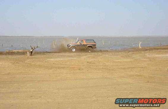 Orange and brown Ford Bronco driving on a dirt path near water, kicking up dust