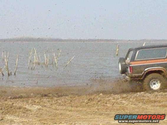 Rear view of black and orange SUV driving through mud near water. Supermotors.net logo visible