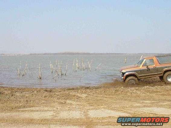 Orange and brown Ford Bronco driving into shallow water from a muddy bank