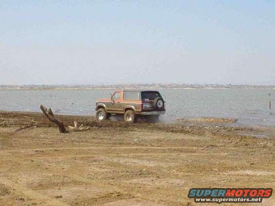 Orange and brown Ford Bronco driving into shallow water on a dirt shore