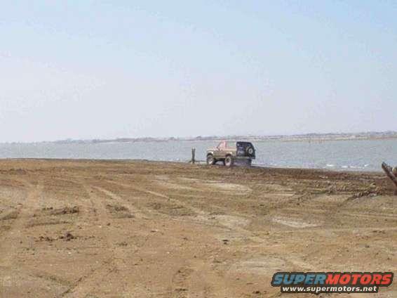 Tan 1980s Ford Bronco with a black spare tire carrier parked on a dirt shore near water
