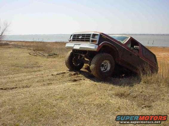 1980s Ford Bronco with lifted suspension and large tires tilted on grassy terrain near water