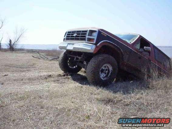 Red and black Ford Bronco tilted on its side in dry grass near a body of water