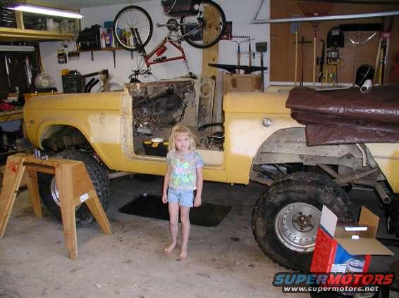 Yellow 1971 Ford Bronco in garage, partially disassembled, with a young girl standing beside it