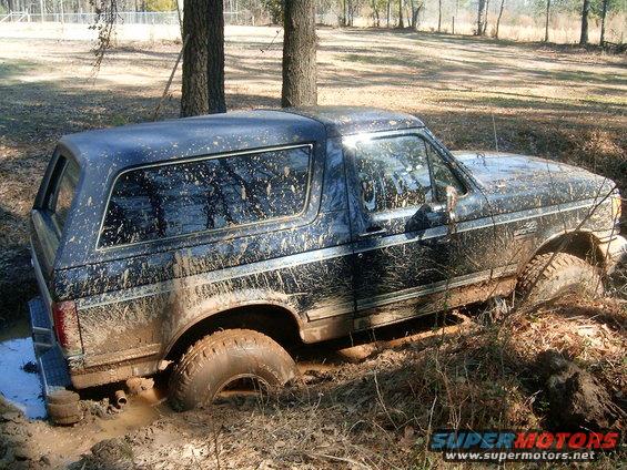 Mud-covered black 1987 Ford Bronco stuck in a muddy ditch, side view
