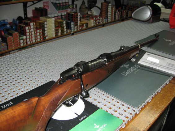 Bolt-action rifle with wooden stock on a counter, next to Swarovski optic brochures