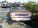 1984 Ford Bronco II - A brown Ford Ranger pickup truck is parked on the side of the road, with a black delivery truck in the background.