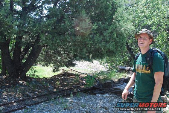 guy in green shirt and hat with backpack standing near some old train tracks and trees