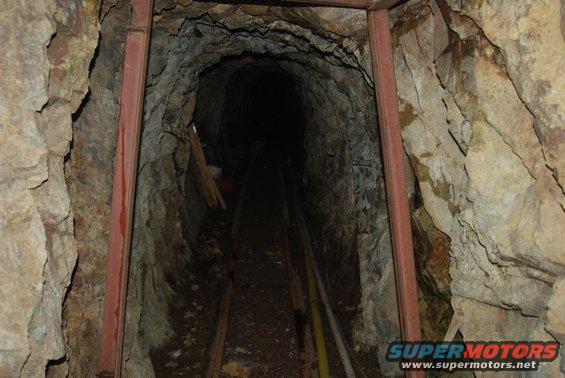 Mine tunnel with wooden supports and metal tracks leading into darkness. Supermotors.net logo in corner