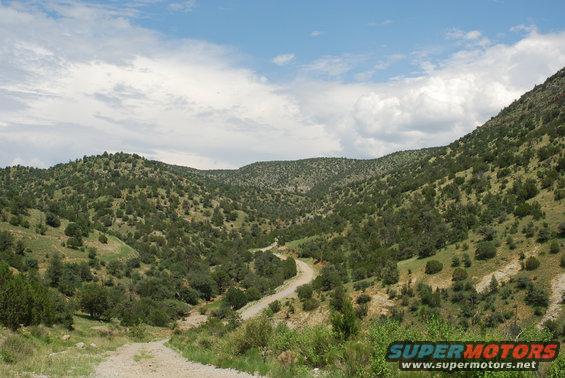 Dirt road winds through a green, hilly valley under a cloudy blue sky